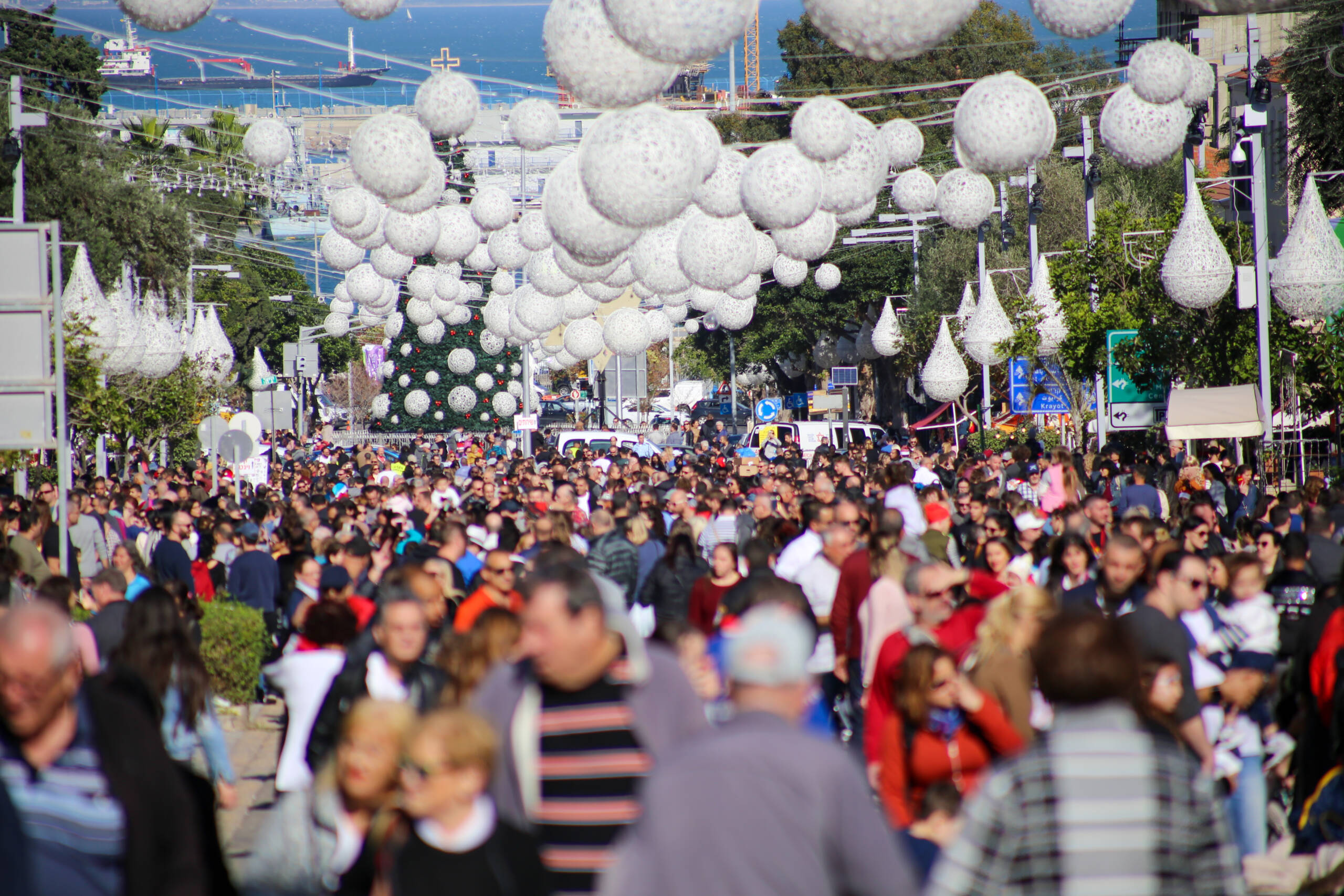 Dense holiday crowd on Ben Gurion Boulevard in Haifa under hanging white woven orbs, with a decorated Christmas tree and the port and bay in the distance